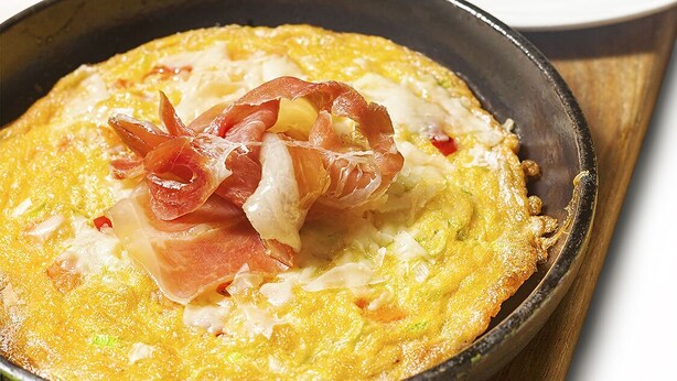 Una sartén con un plato, posiblemente una tortilla, adornada con tocino y tomates, sobre una tabla de madera.