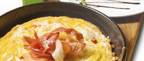 Een koekenpan met een gerecht, mogelijk een omelet, versierd met spek en tomaten, op een houten plank.