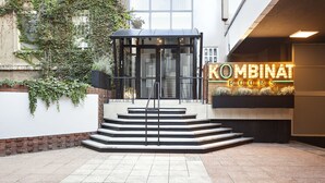 Entrada de un edificio con puerta de vidrio, escalones que conducen a ella y un letrero de neón que dice "KOMBINAT ROOMS".