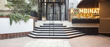 A building entrance with a glass door, steps leading up to it, and a neon sign reading "KOMBINAT ROOMS".