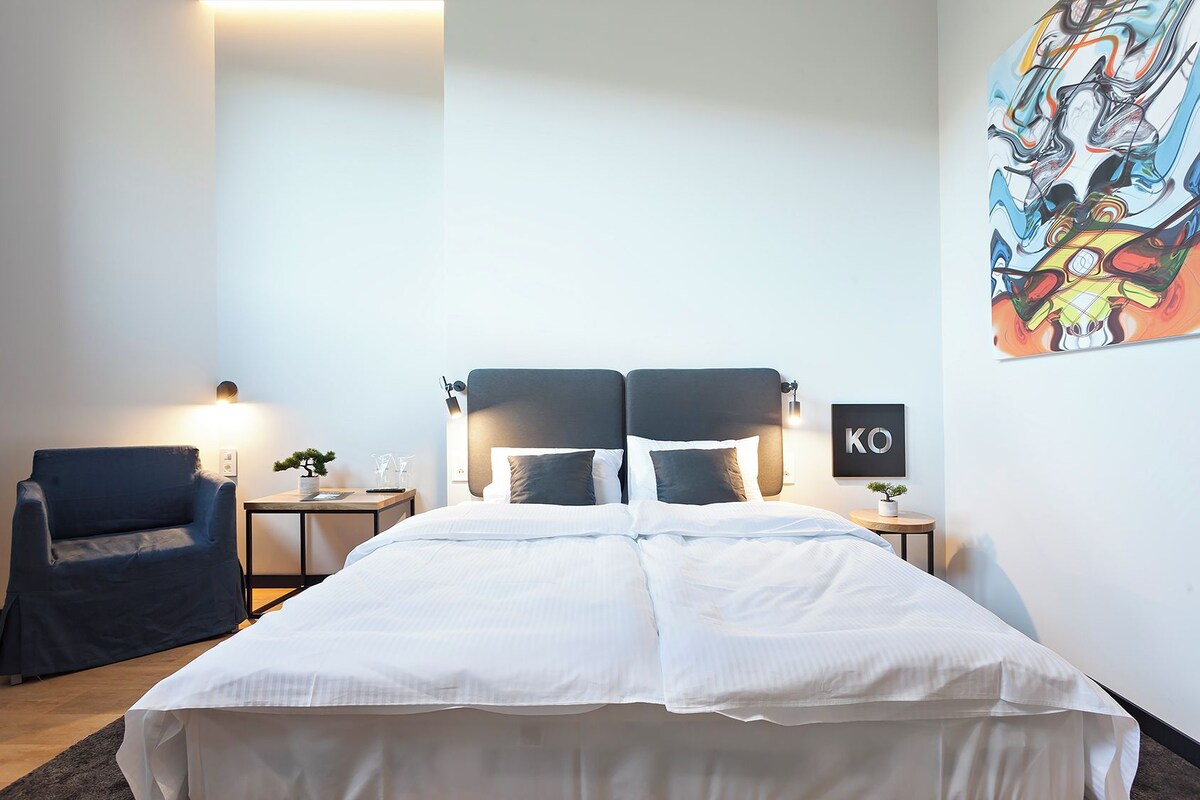 Una habitación de hotel moderna con una cama grande, una silla, una mesita de noche y una obra de arte colgada en la pared.