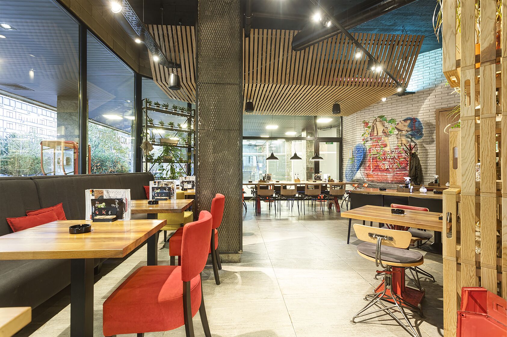 A modern cafe with wooden tables and red chairs, a mural on the wall, and large windows overlooking a cityscape.