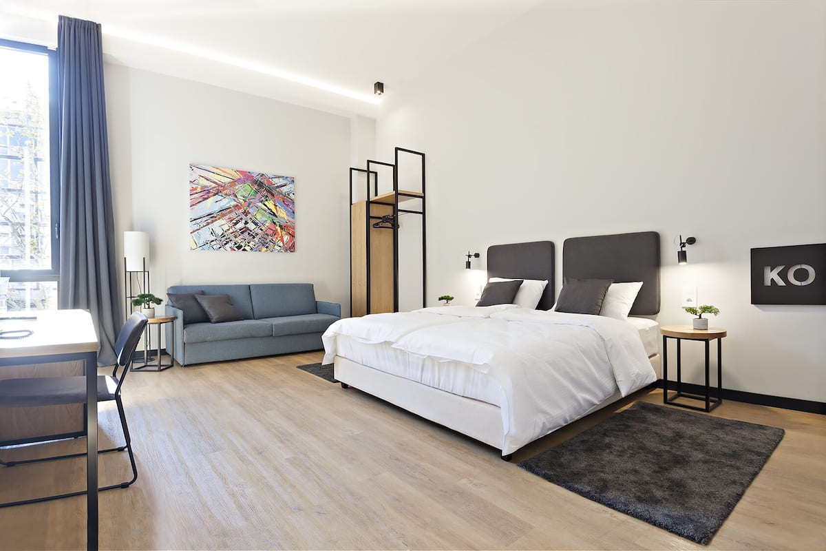 Een moderne hotelkamer met een groot bed, een bureau, een bank, een kleurrijk abstract schilderij en een 'KO' bord op de muur.
