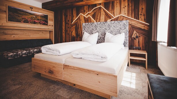 A wooden-framed bed with white linens, a headboard with a geometric pattern, and a small bedside table. - Gschoadwirt (Sankt Aegyd am Neuwalde)