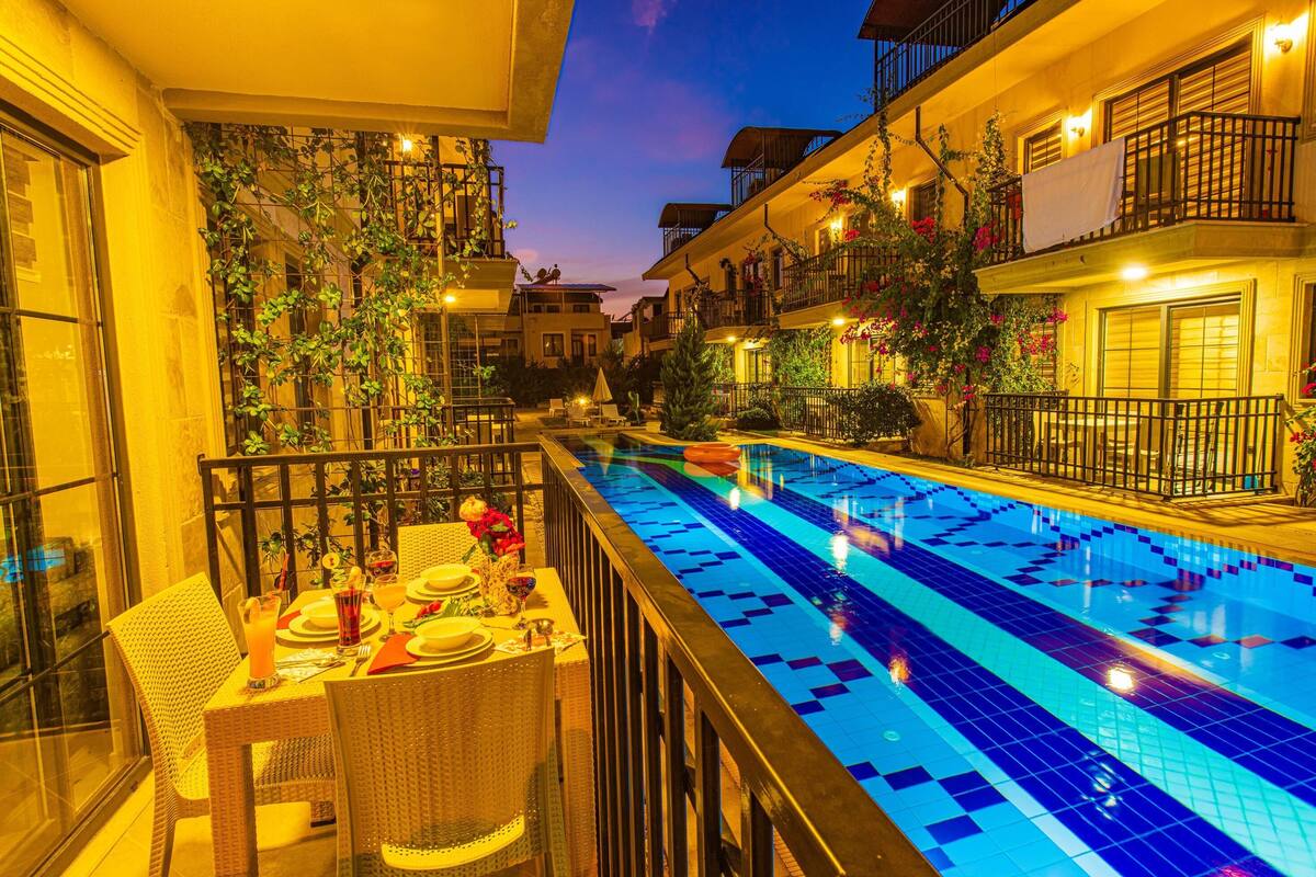 Un espace repas aménagé au bord de la piscine, avec une table dressée pour deux personnes, entouré d’une rambarde, et une piscine dont le fond est recouvert de carreaux bleus.