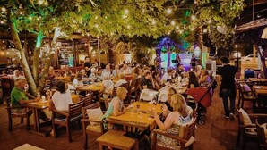 An outdoor dining area with tables and chairs under trees, surrounded by buildings.