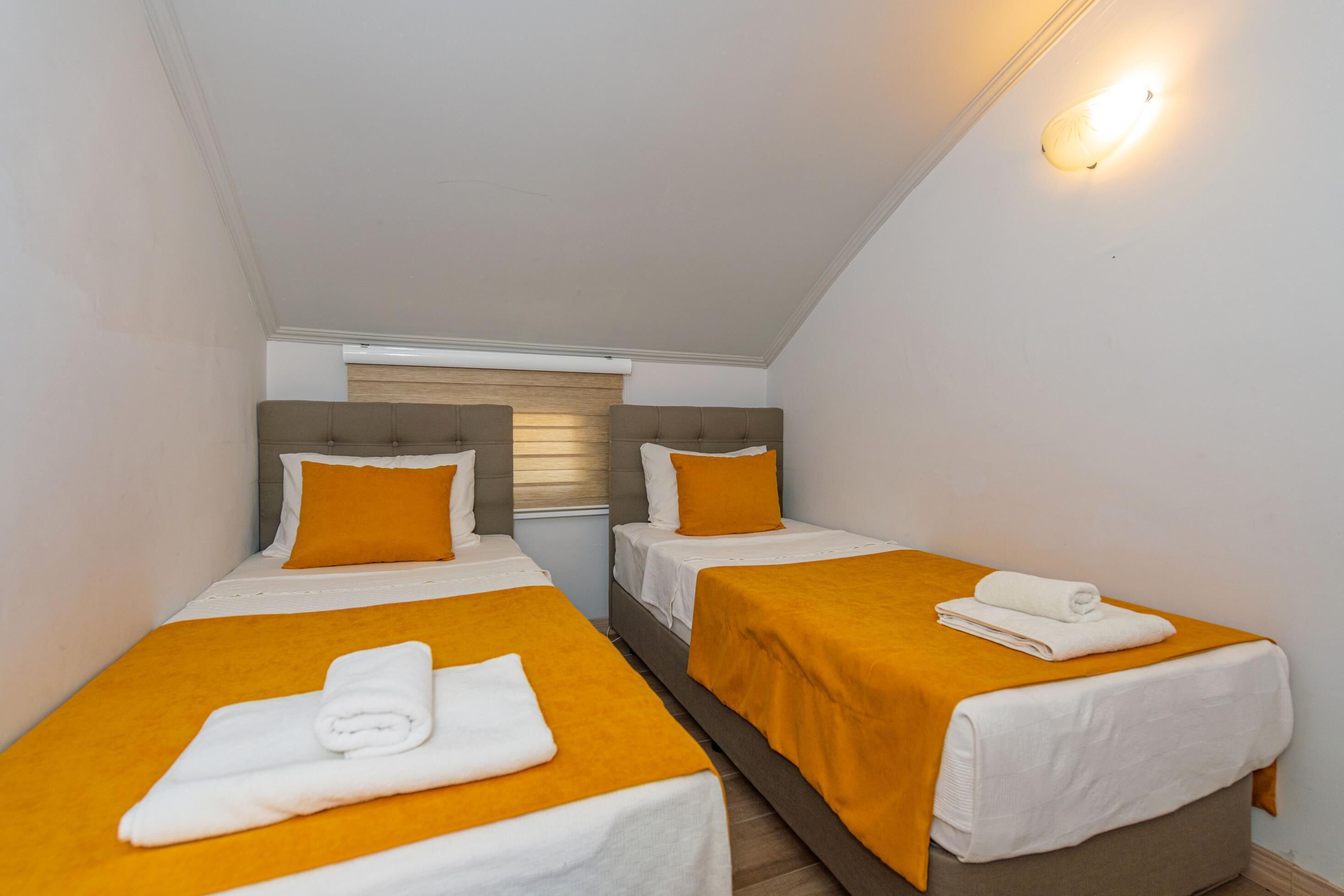 A small, well-lit room with two single beds, each with a white and orange bedspread, and a neatly folded towel on each bed.