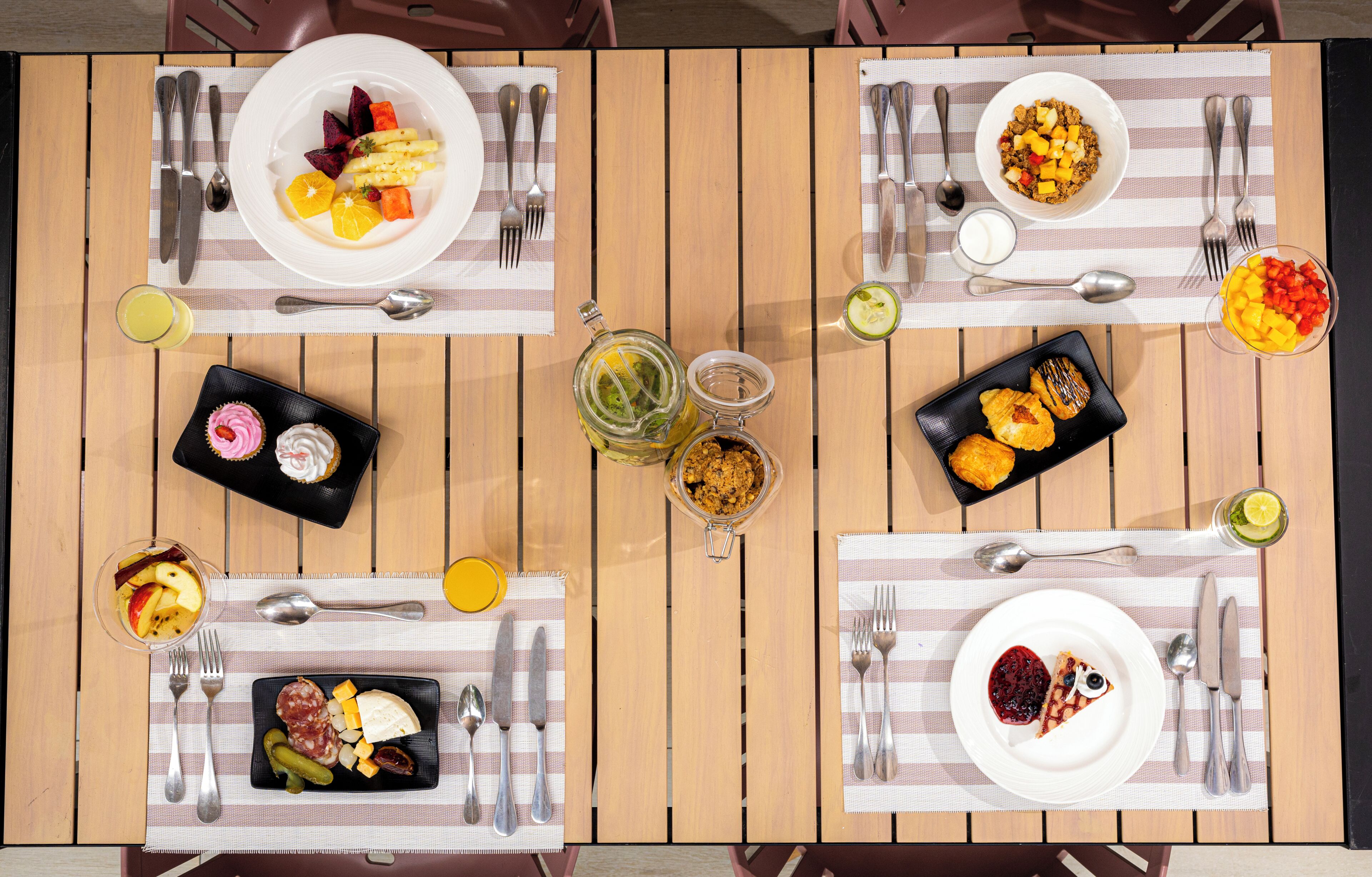 A wooden table with four sets of dining utensils, plates with various foods, and a glass of juice.