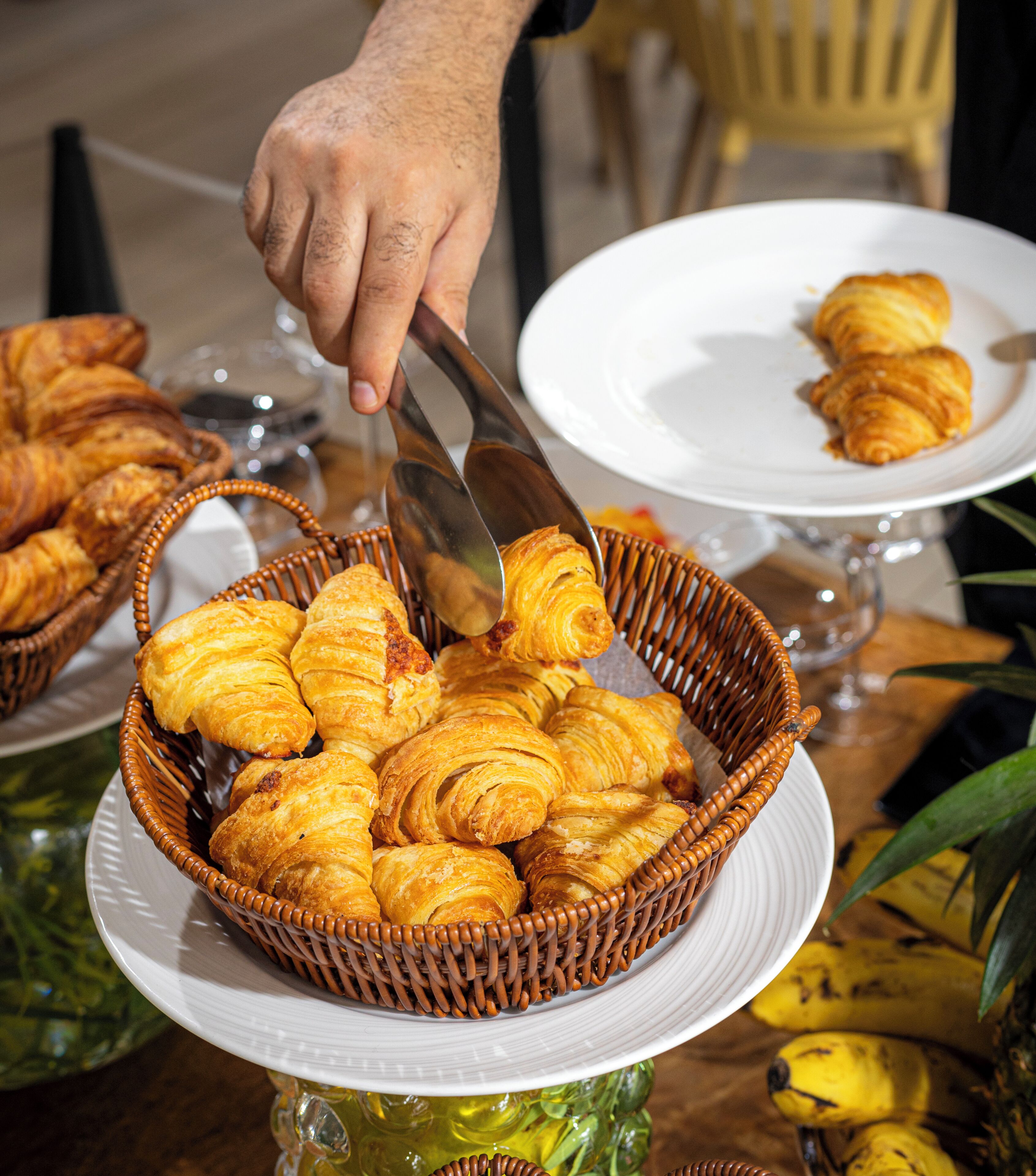 A basket of croissants on a white plate, a hand holding a knife, and a glass of water on a table.