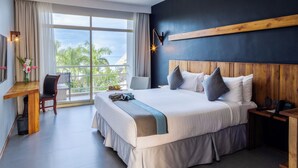 A hotel room with a large bed, a desk with a chair, a nightstand, and a view of palm trees through the window.