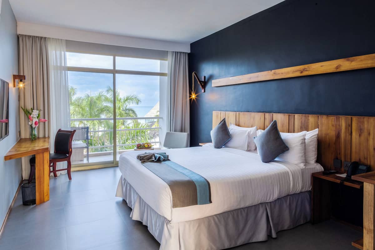 A hotel room with a large bed, a desk with a chair, a nightstand, and a view of palm trees through the window.