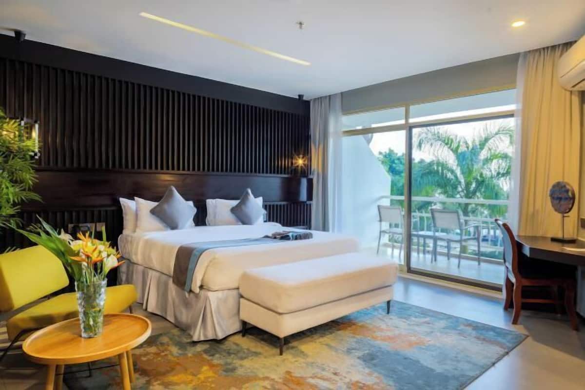 A modern hotel room with a large bed, a desk, a chair, a small table with a vase, and a view of the outdoors.