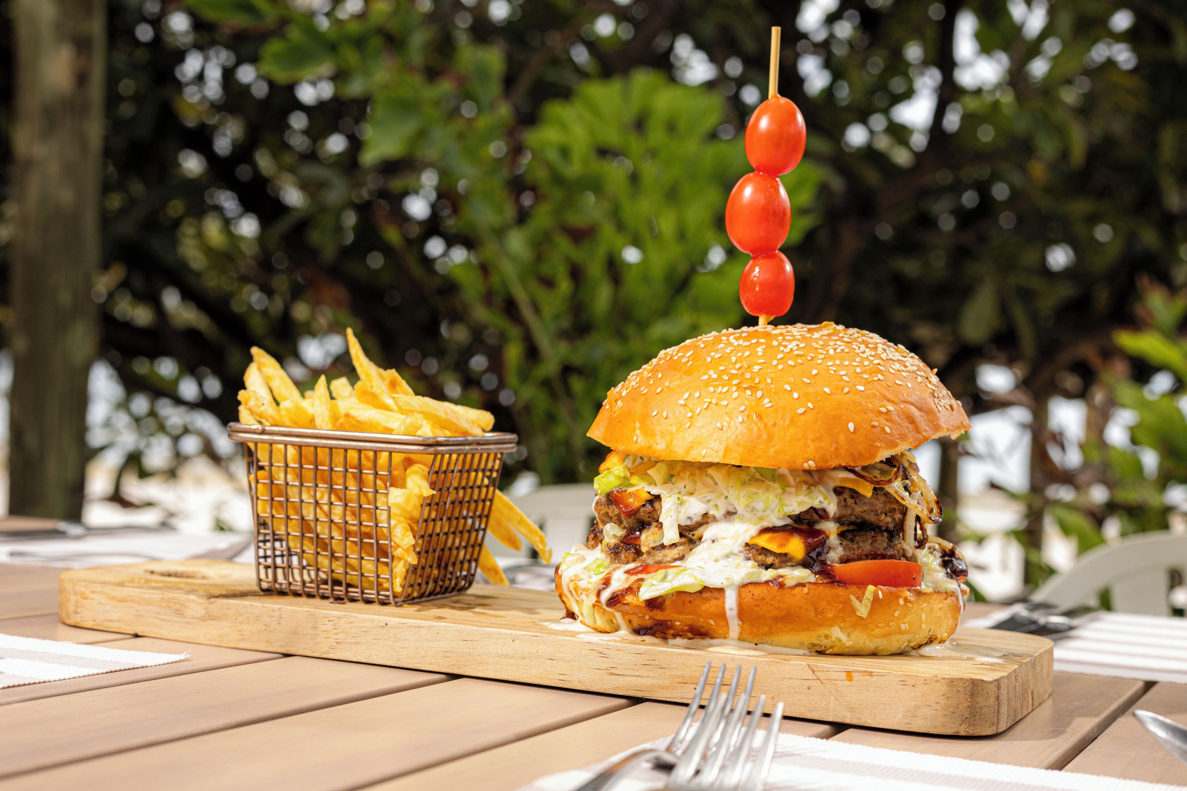A burger with a skewer of tomatoes on top, a basket of fries, and a wooden cutting board on a table.