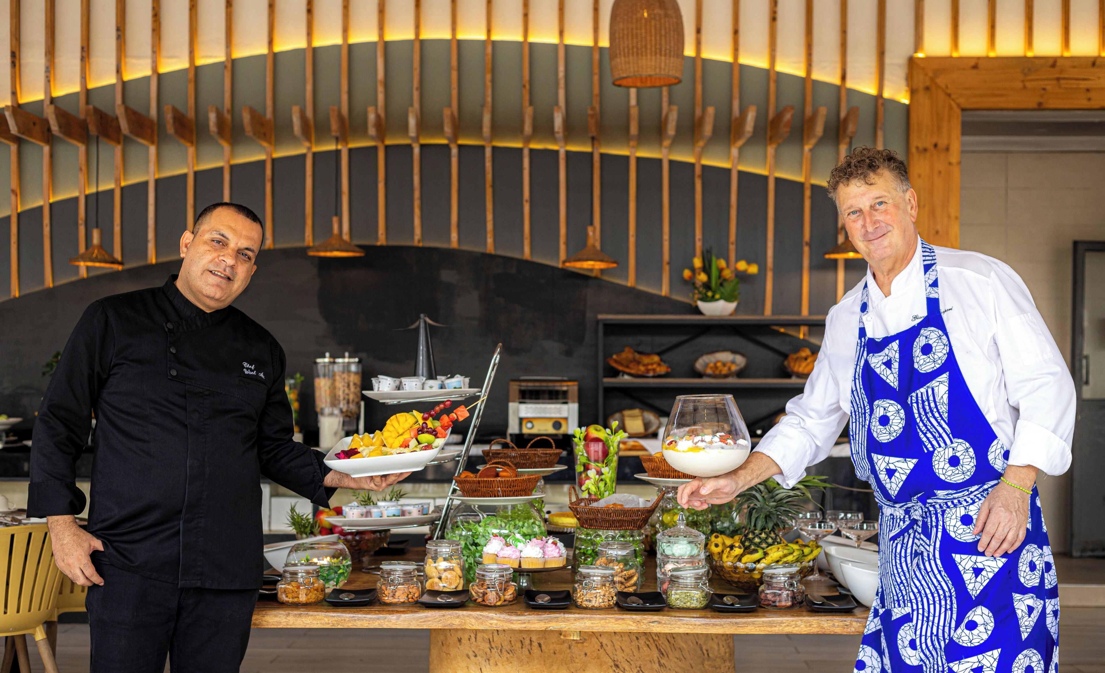 Twee koks presenteren gerechten bij een buffet met diverse etenswaren en een moderne keuken op de achtergrond.
