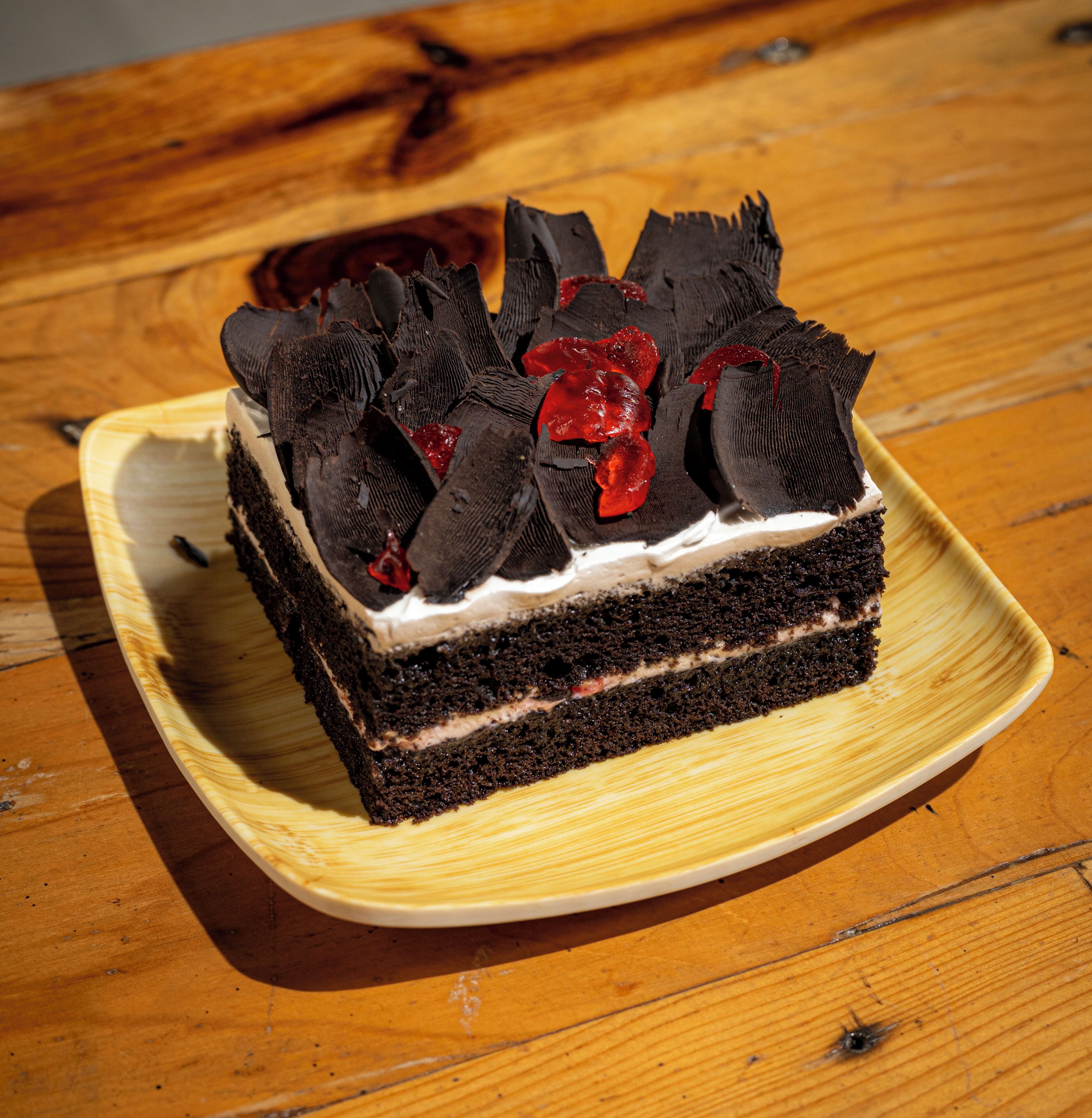 A slice of chocolate cake with whipped cream and red fruit on a square plate on a wooden table.