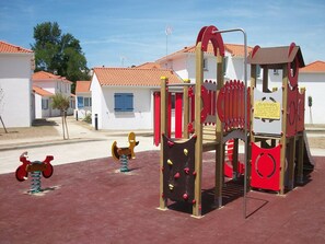 Un terrain de jeux avec des structures d’escalade et un toboggan, entouré de bâtiments blancs aux portes rouges.