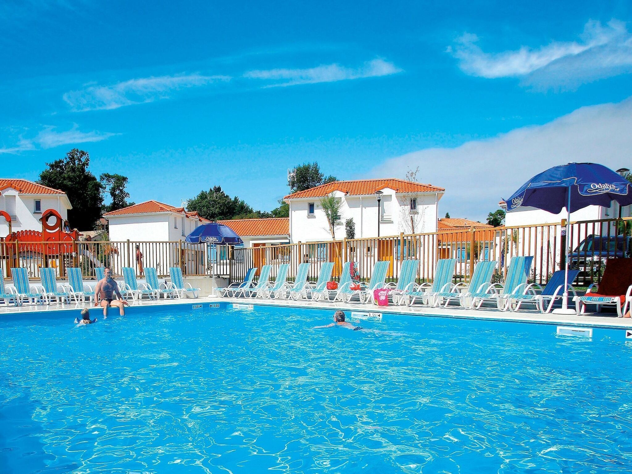 Ein Swimmingpool mit Liegestühlen und Sonnenschirmen, umgeben von Gebäuden mit roten Ziegeldächern.