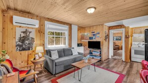 Une pièce confortable, aménagée dans le style d’un chalet, avec un canapé gris, un plafond en bois et un coin cuisine équipé d’une cuisinière et d’un réfrigérateur.