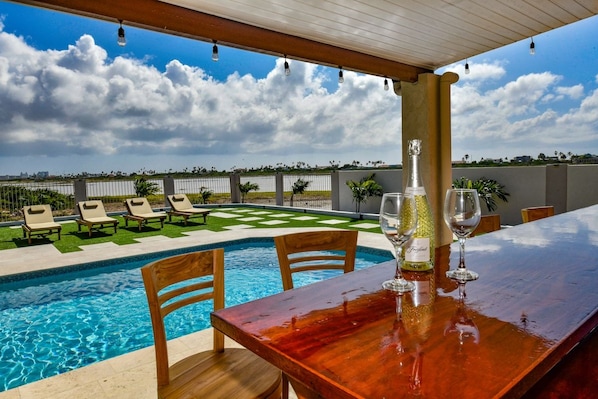 Un bar junto a la piscina con una mesa y sillas de madera, una botella de vino y copas sobre la mesa.