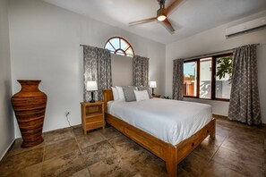 Un dormitorio con cama, mesitas de noche, un jarrón grande, un ventilador de techo y una ventana con cortinas.