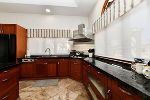 A modern kitchen with wooden cabinets, a black refrigerator, a stainless steel range hood, and a window with a striped blind. - Pool Oasis: 3BR Home w/ Patio near Arashi Beach (Noord)