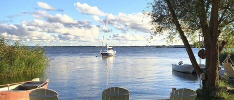 Three Adirondack chairs facing a calm lake with boats and a sailboat in the distance.