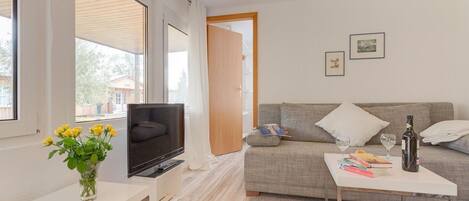 Una sala de estar moderna con un televisor de pantalla plana, un sofá, una mesa de centro con libros y una botella de vino, y un jarrón con flores.