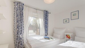 A bedroom with two beds, a window with curtains, a bedside table with books, and a wall with framed pictures.