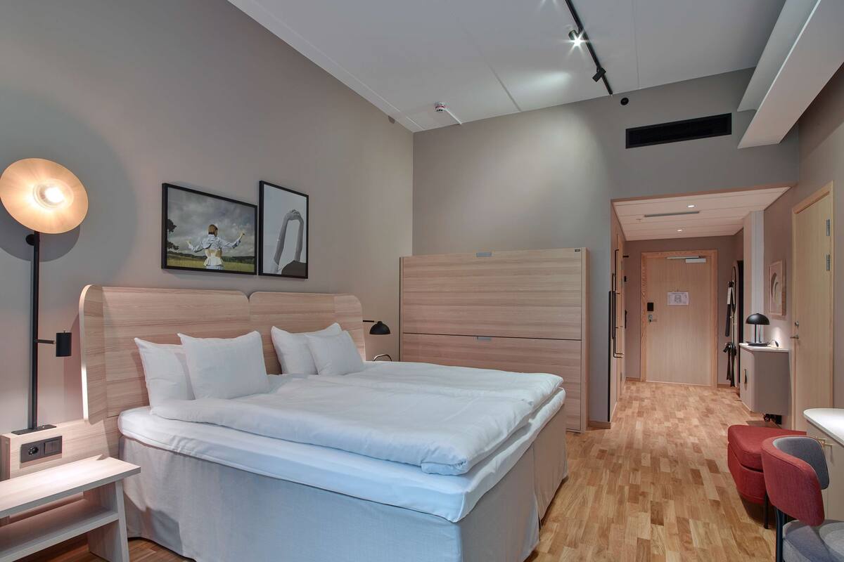 A modern hotel room with a large bed, bedside tables, a desk, and a chair. There is a hallway leading to other rooms.