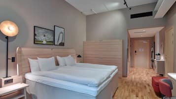 Una habitación de hotel moderna con una cama grande, mesitas de noche, un escritorio y una silla. Se ve un pasillo que conduce a otras habitaciones.