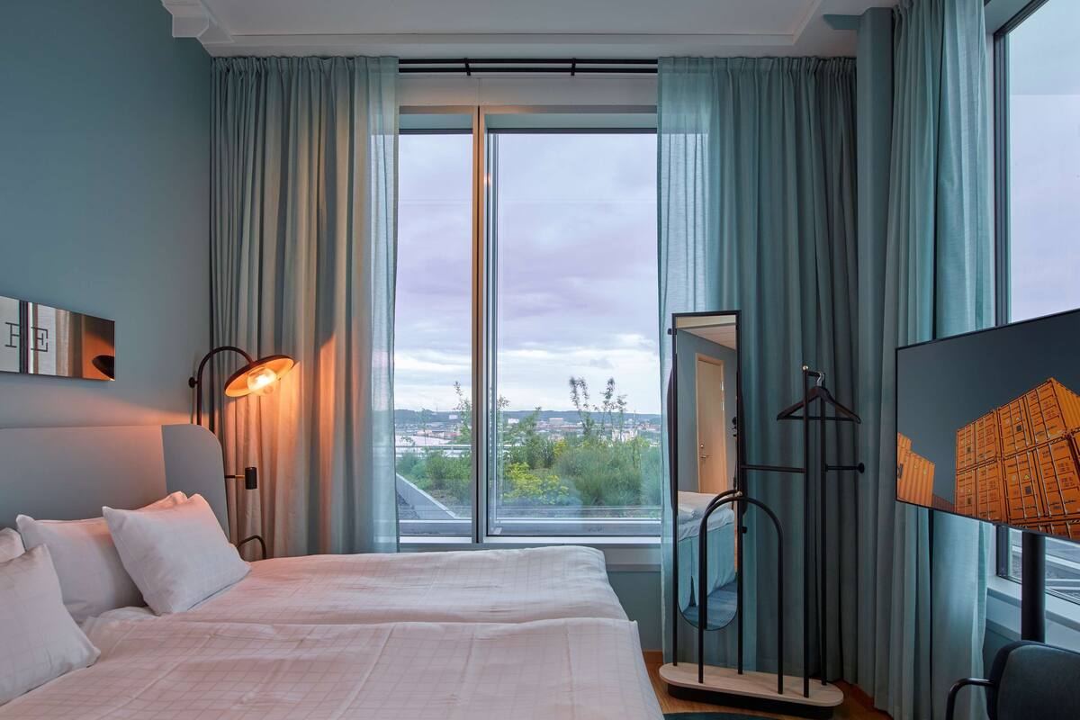 A hotel room with a large bed, a desk, a chair, a TV, and a view of the city through large windows.