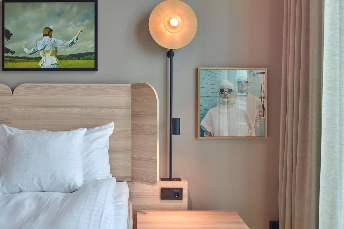 A hotel room with a bed, a nightstand, a wall-mounted lamp, and two framed pictures.