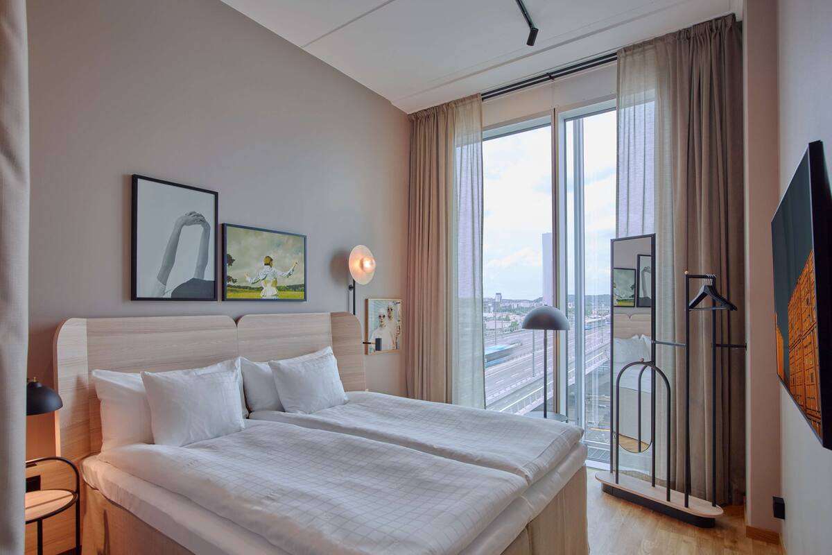 A hotel room with a large bed, a desk, and a view of the city through large windows.