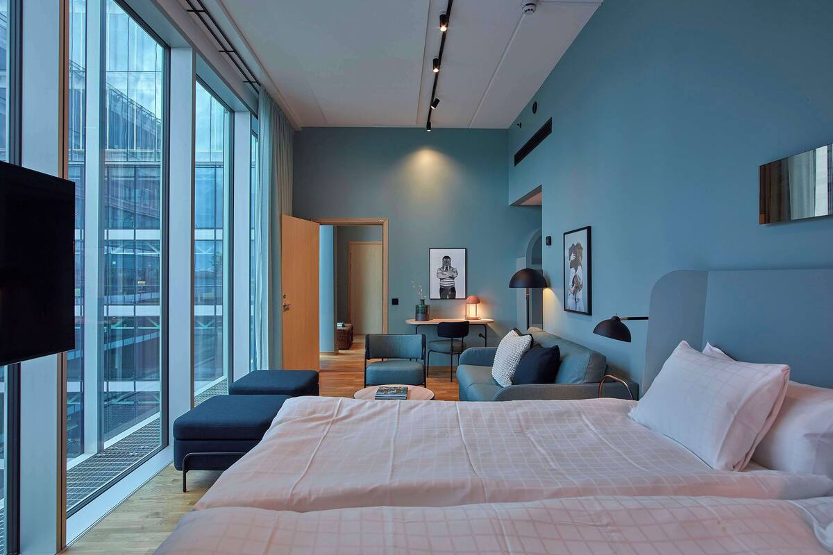 A modern hotel room with a large bed, a desk, and a sofa. There is a TV mounted on the wall and a window providing a view of the city.