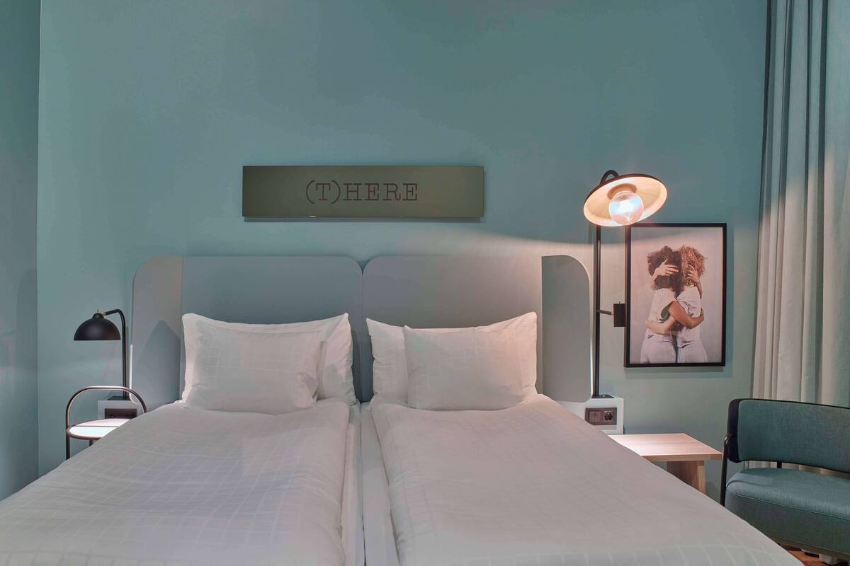 A double bed with white linens, a bedside table with a lamp, a chair, and a framed picture on the wall.