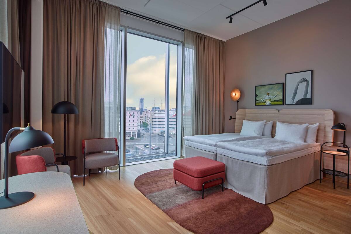 A modern hotel room with a large bed, a desk, a chair, a red ottoman, and a view of the city through floor-to-ceiling windows.