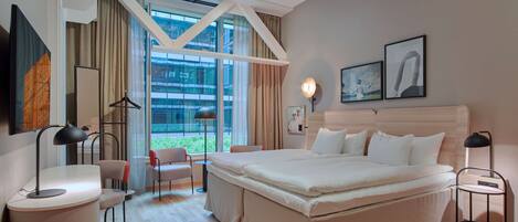 A modern hotel room with a large bed, a desk, a chair, and a view of the outdoors.