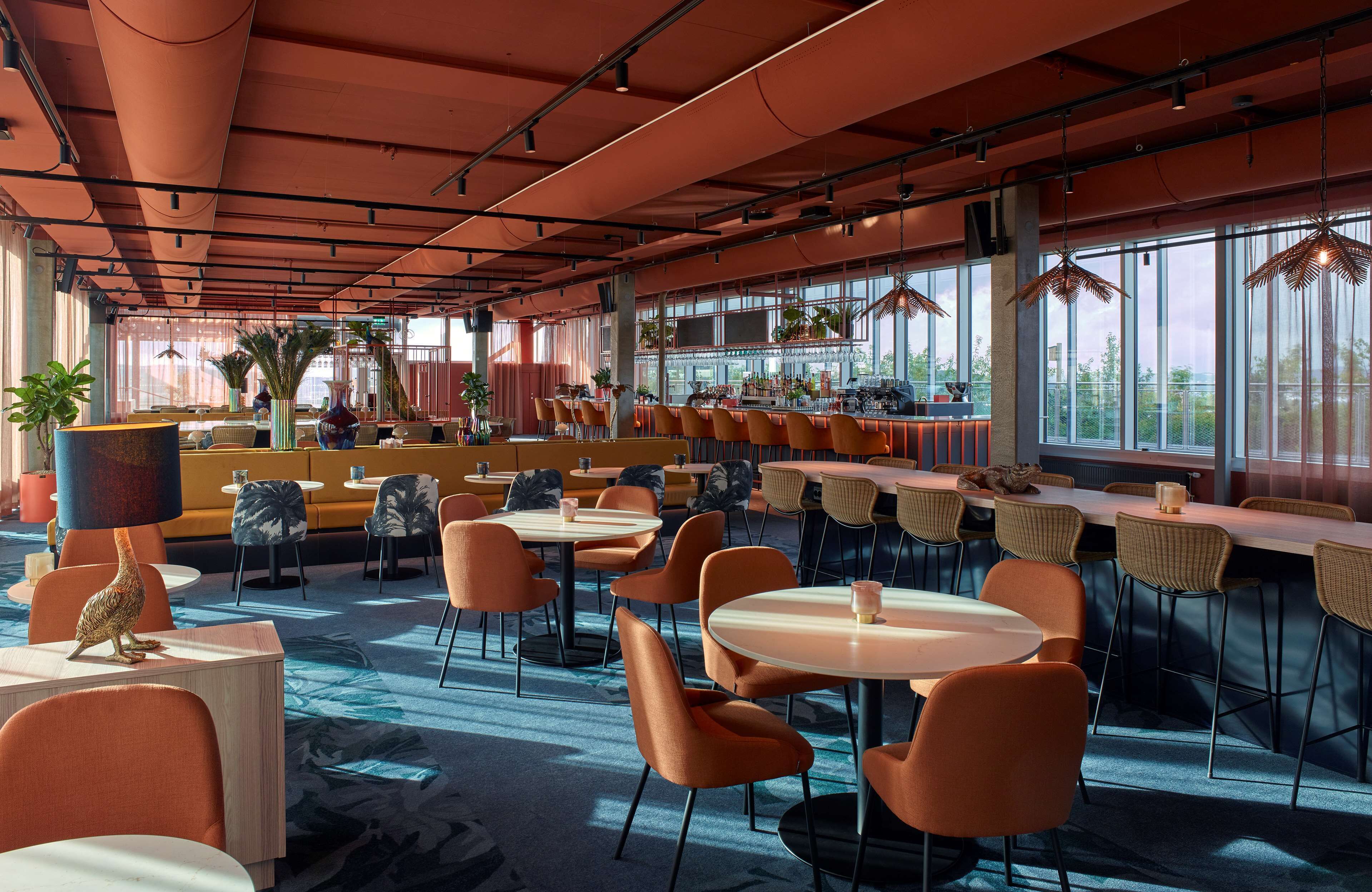 En rymlig restaurang med högt i tak, stora fönster och ett varierat utbud av sittplatser, inklusive bord och avskilda sittgrupper.