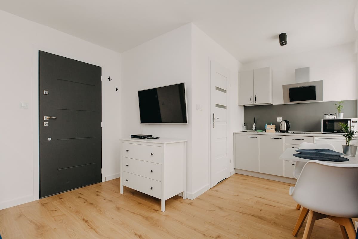 A modern kitchen with white cabinets, a black door, a flat-screen TV, and a dining area with a white table and chair.