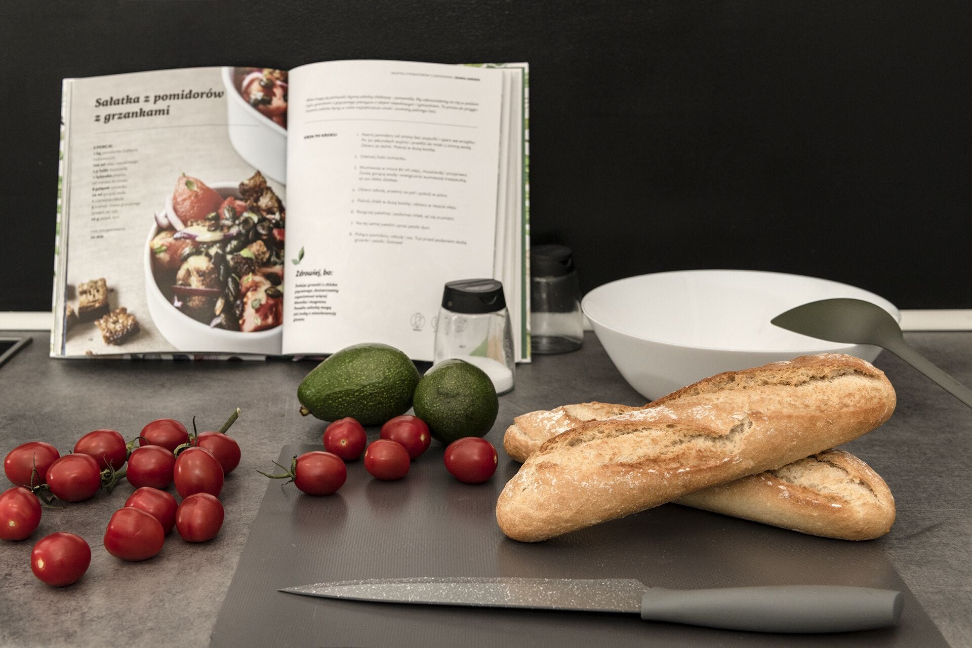 A cookbook open on a table with ingredients like tomatoes, avocados, and bread, suggesting a cooking theme.
