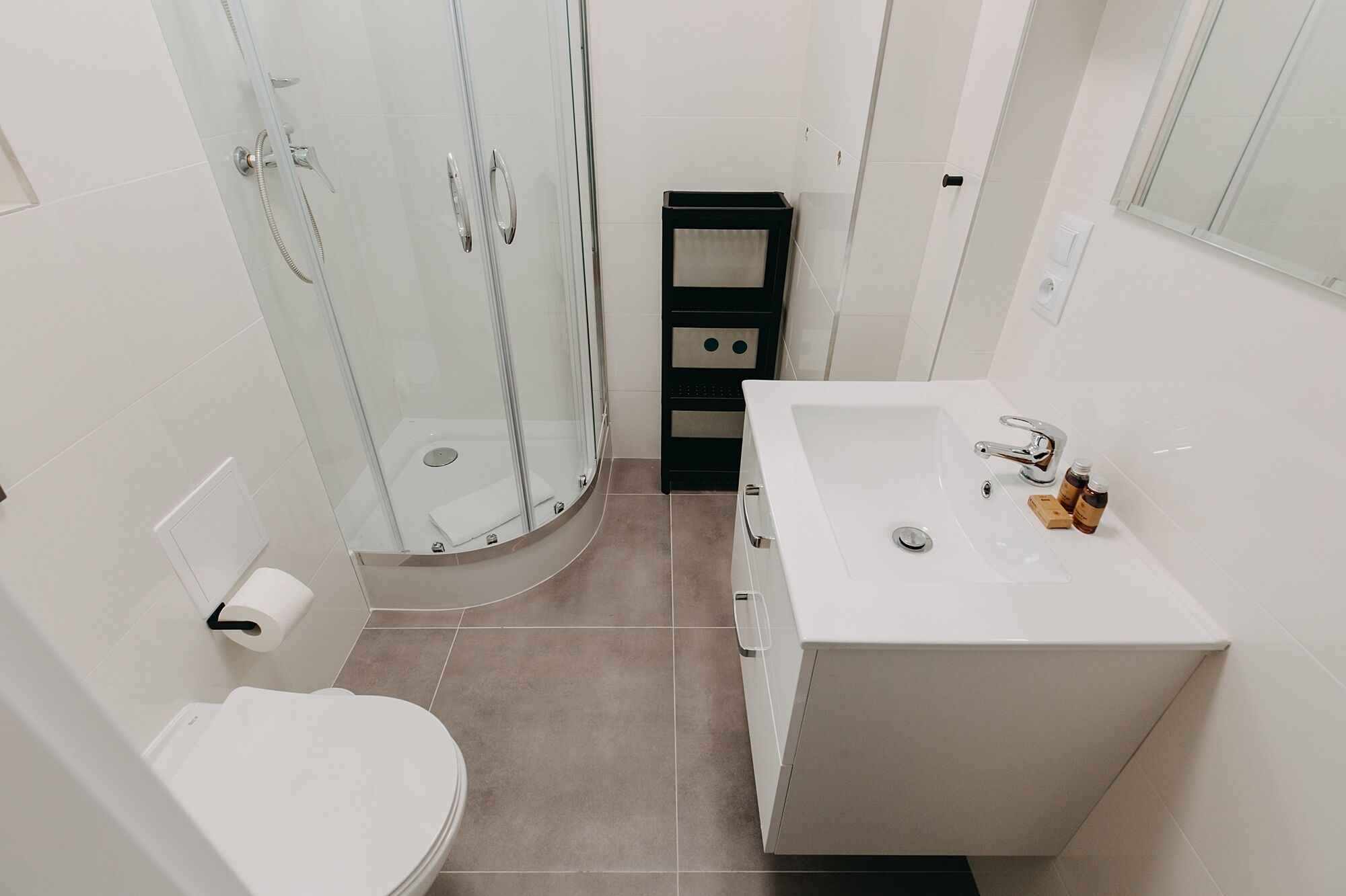 A modern bathroom with a glass shower enclosure, a white toilet, a sink with a mirror, and a black storage unit.
