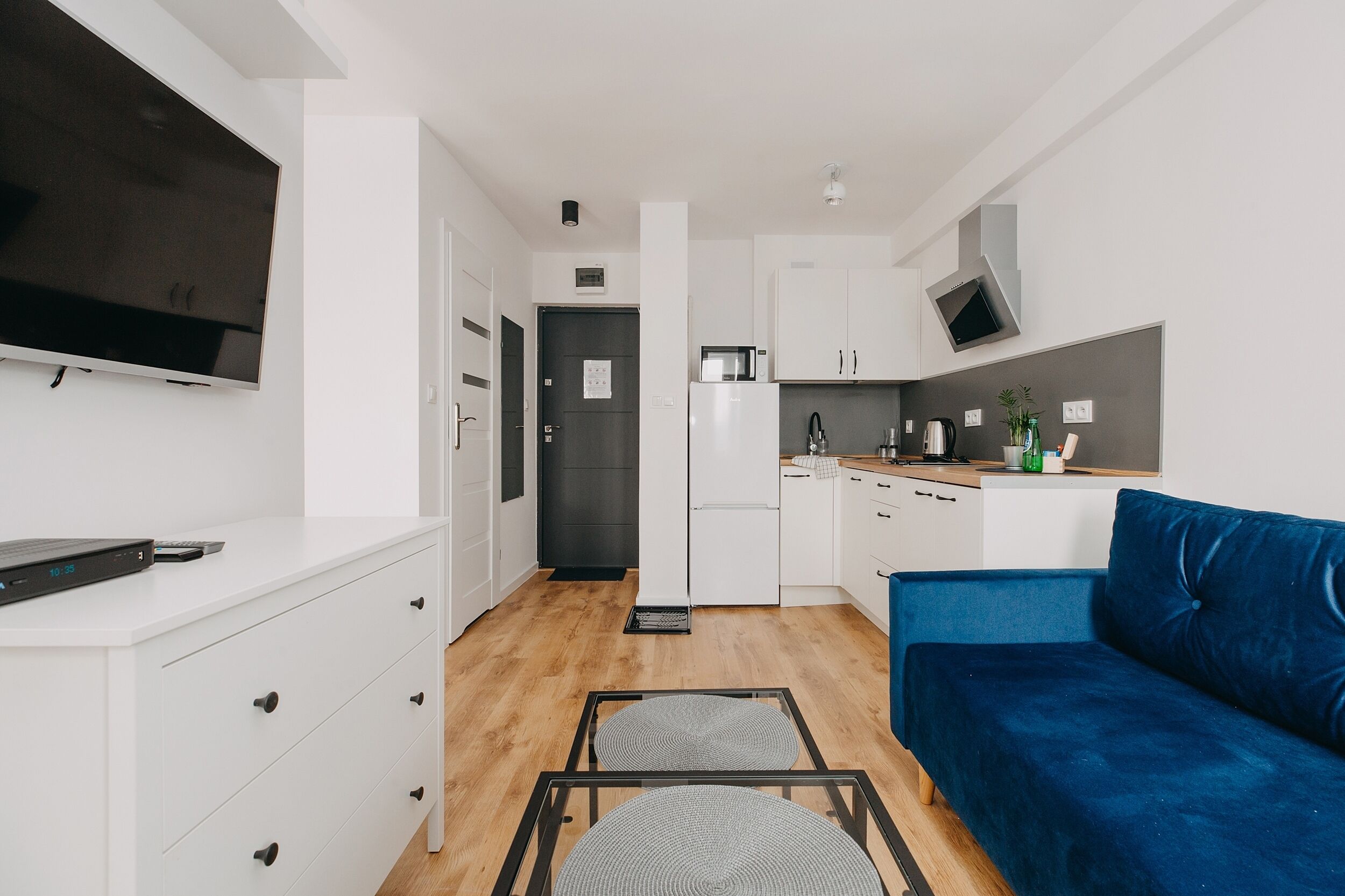 Un espacio habitable moderno y funcional con kitchenette, un televisor de pantalla plana montado en la pared, un sofá azul y una pequeña mesa de comedor con sillas.
