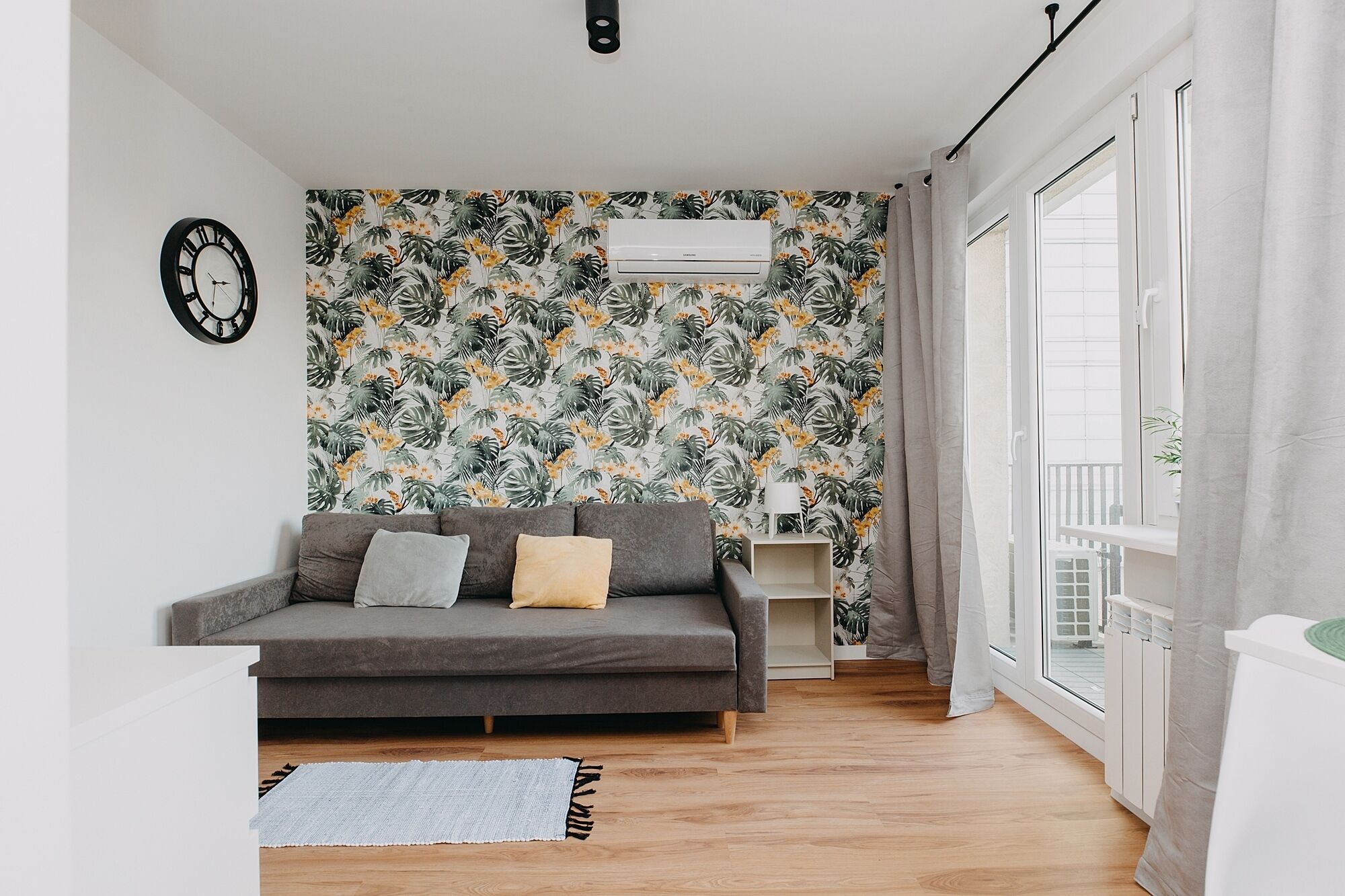 Ein Wohnzimmer mit grauem Sofa, strukturierten Tapeten, einer Uhr und einem Fenster mit Vorhängen.