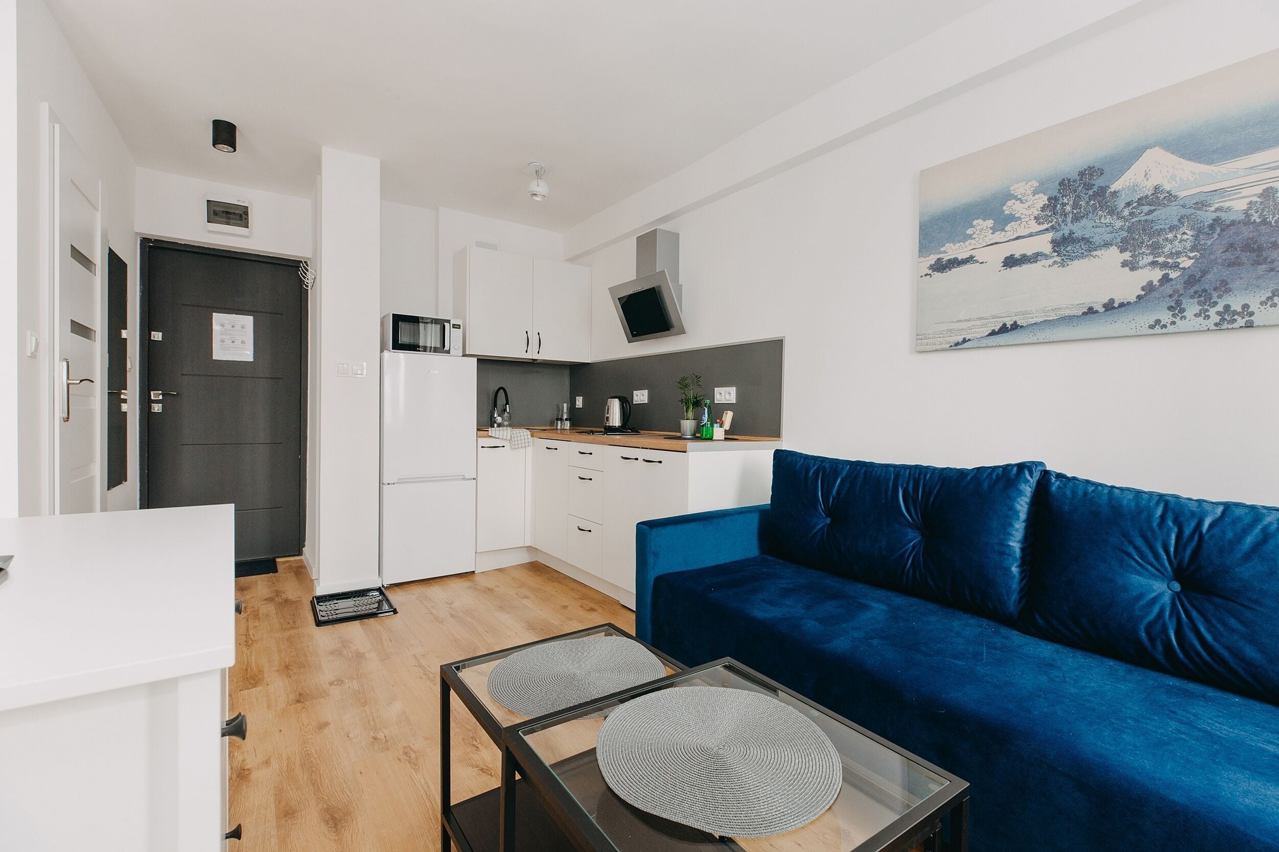Una cocina moderna con armarios blancos, un microondas negro y un fregadero. Hay un sofá azul con un cojín y una pequeña mesa con un mantel individual. Un cuadro de un paisaje montañoso está en la pared.