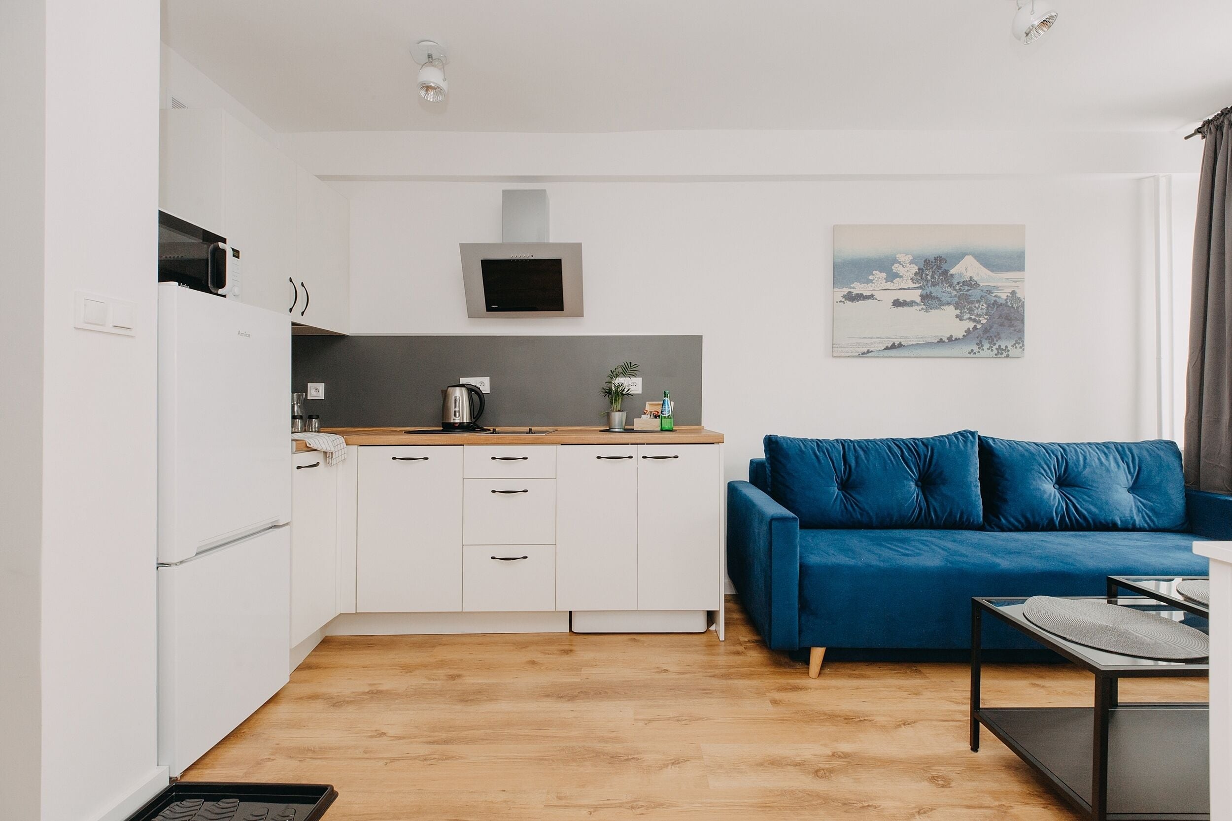 A compact kitchen with white cabinets, a microwave, and a kettle on the counter. There's a blue sofa and a small table with a tray.
