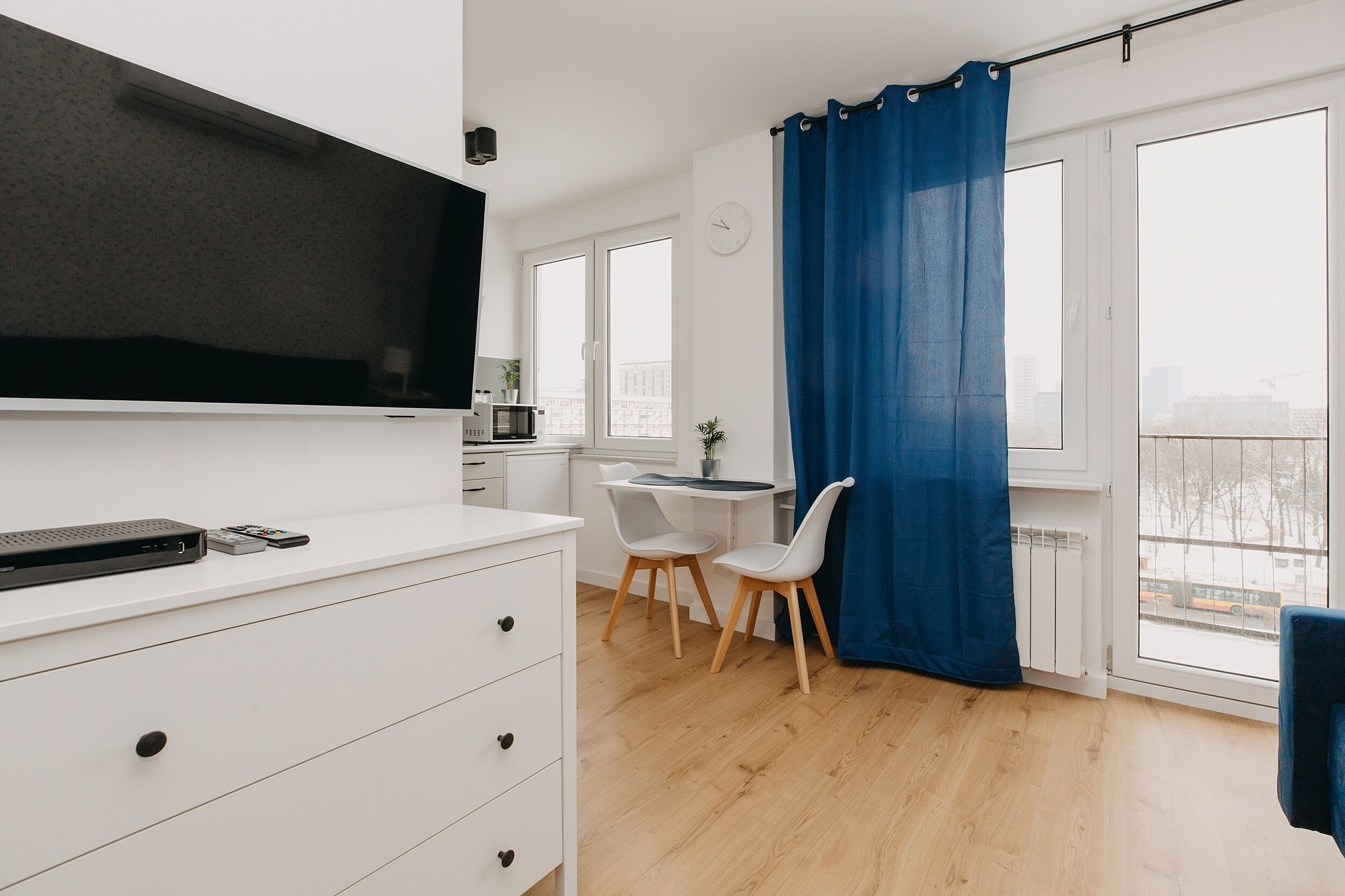 A modern apartment with a flat-screen TV, a dining area with two chairs, a large window with a blue curtain, and a view of the city.