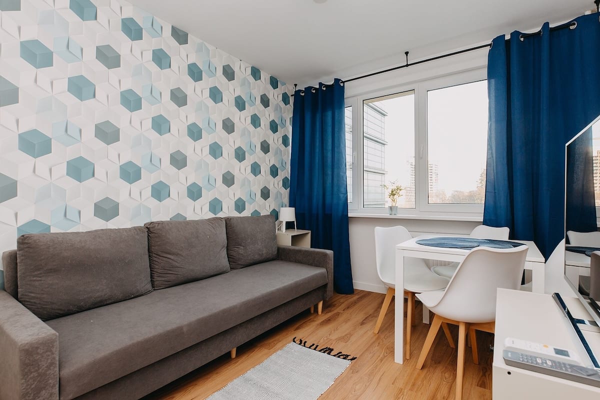 A modern living room with a grey sofa, white chairs, a small table, and a window with blue curtains.