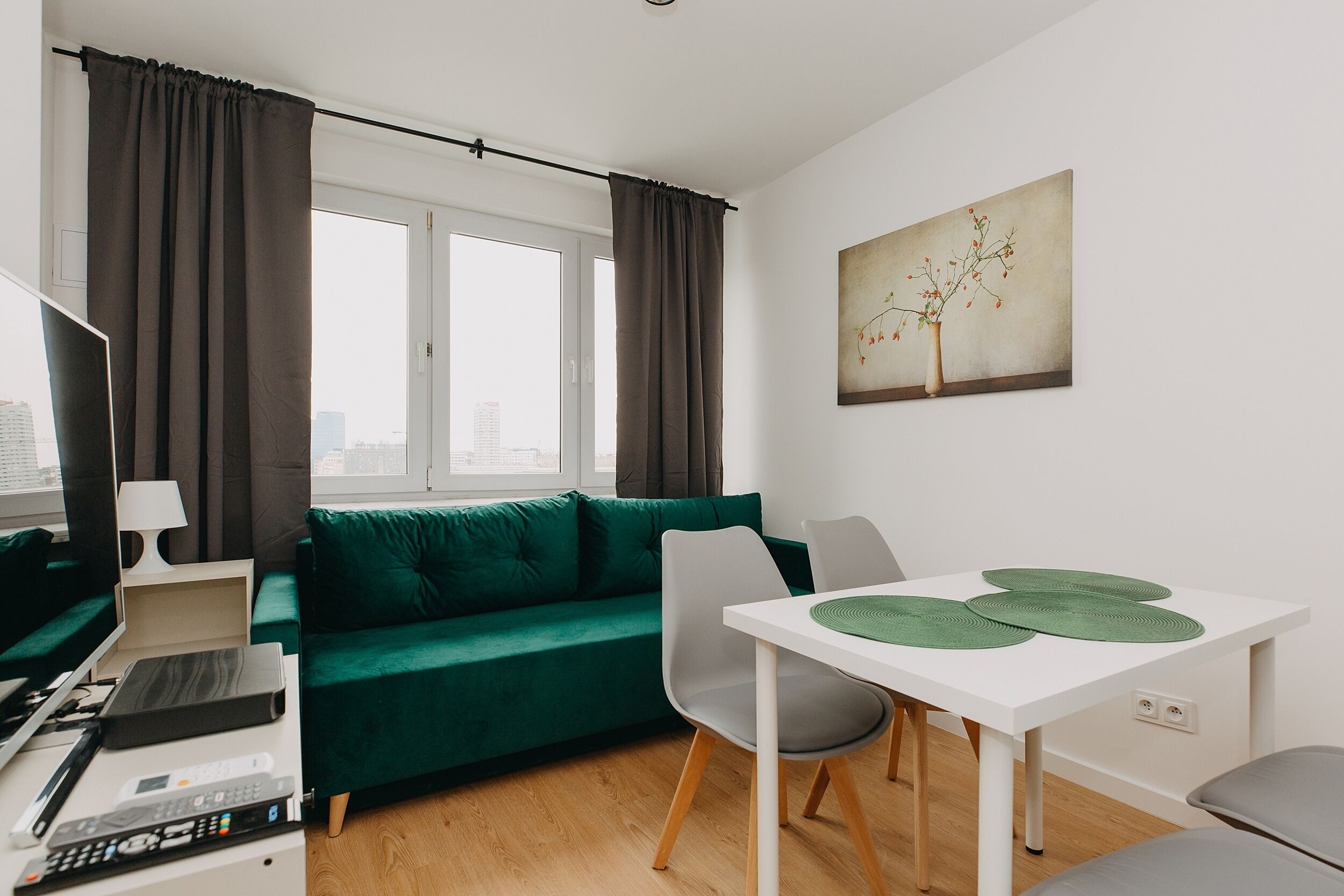 Una sala de estar moderna con un sofá verde, una mesa de comedor blanca con individuales verdes y vistas a la ciudad a través de grandes ventanales.