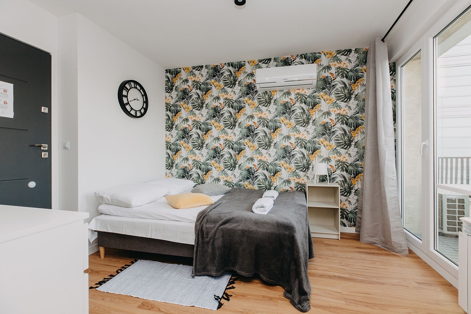 Un dormitorio con cama, una manta gris, almohada blanca, reloj, aire acondicionado y ventana con cortinas.