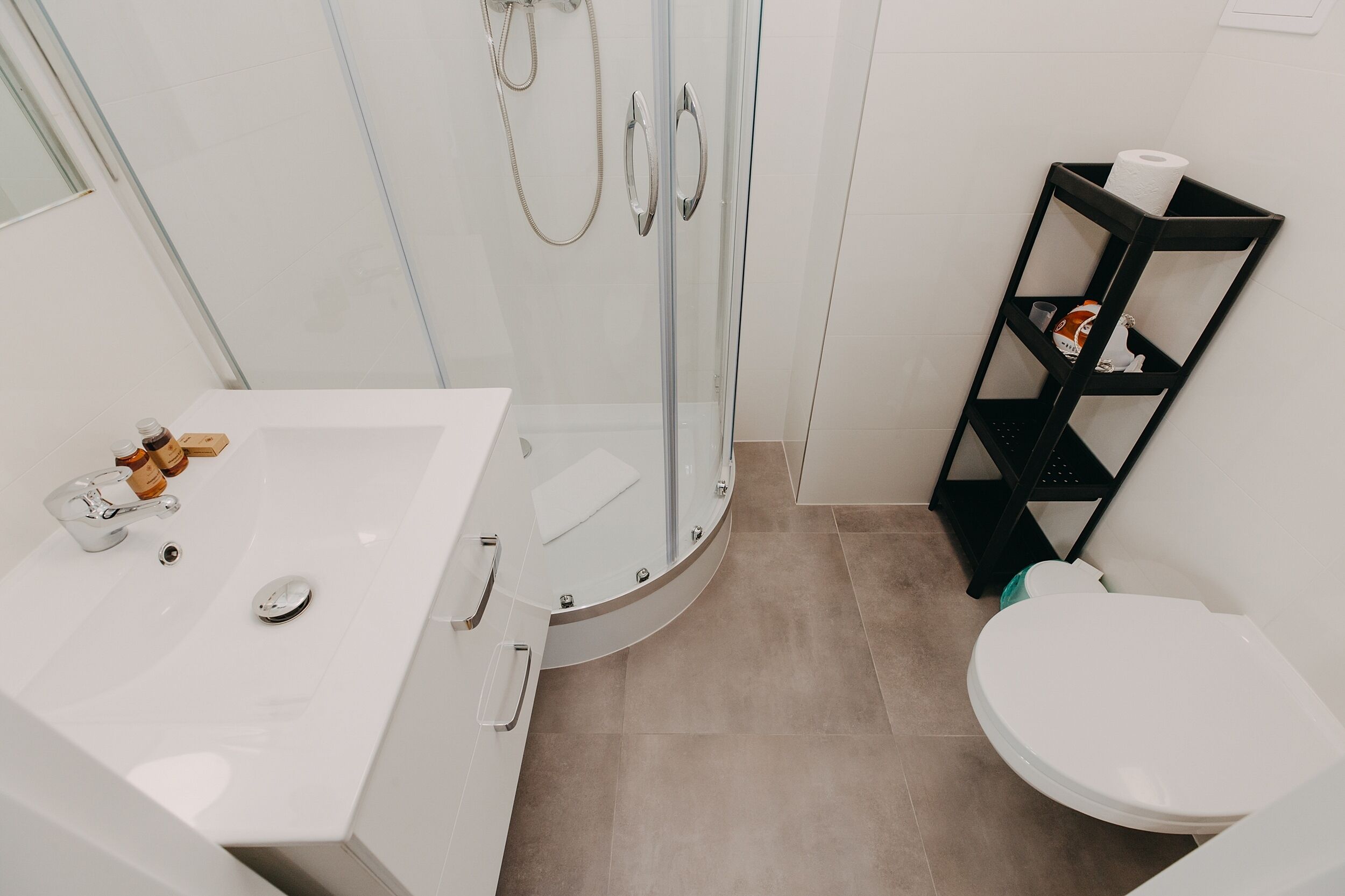 A modern bathroom with a glass-enclosed shower, a white sink with toiletries, and a black shelf with a toilet paper roll.